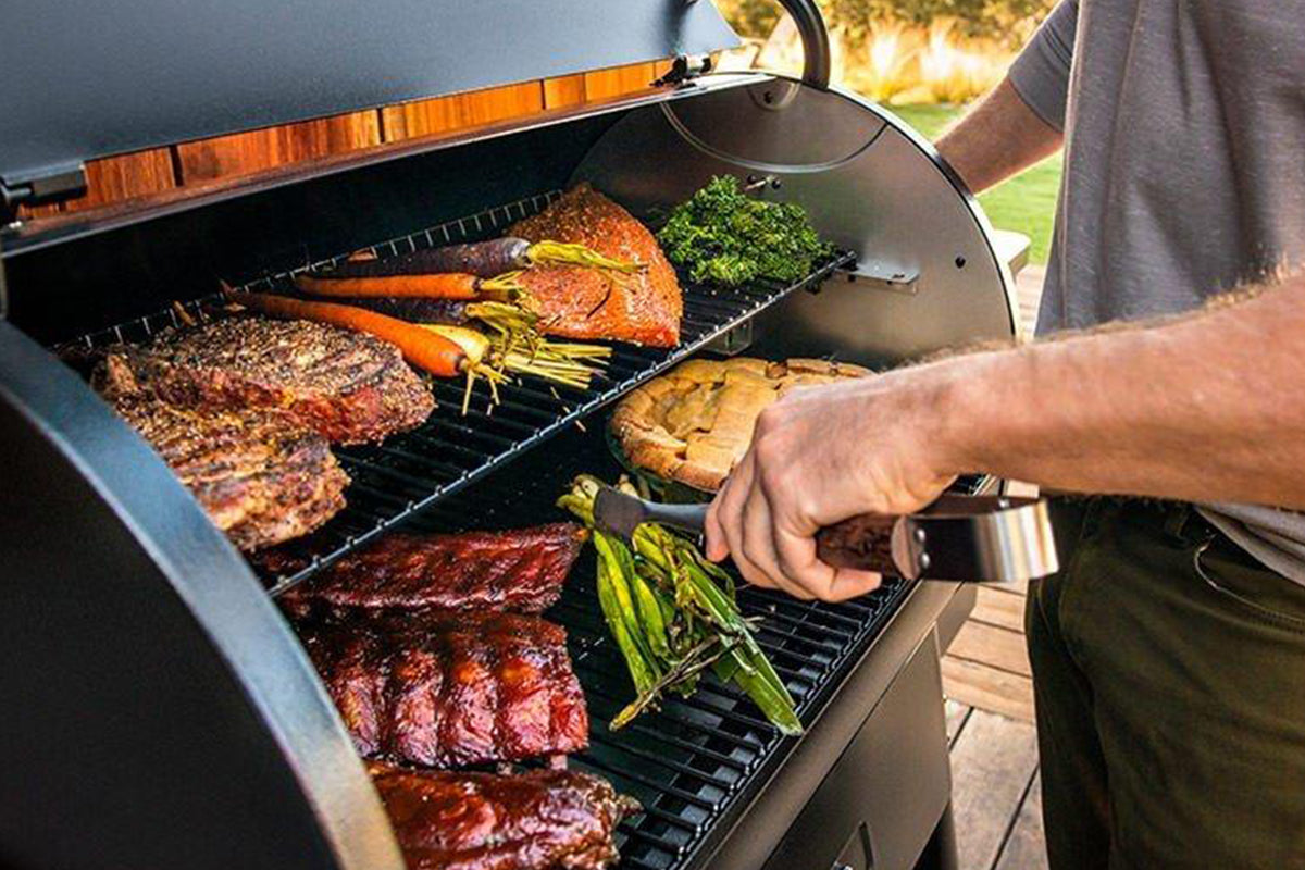Person using tongs to turn asparagus on a Traeger grill filled with meat and vegetables