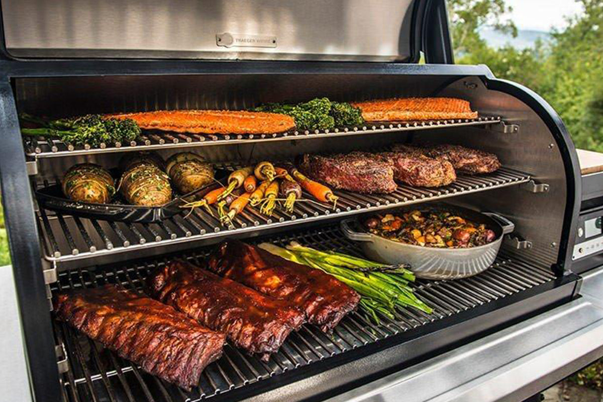 Open Traeger grill with three cooking racks holding salmon, meat and vegetables
