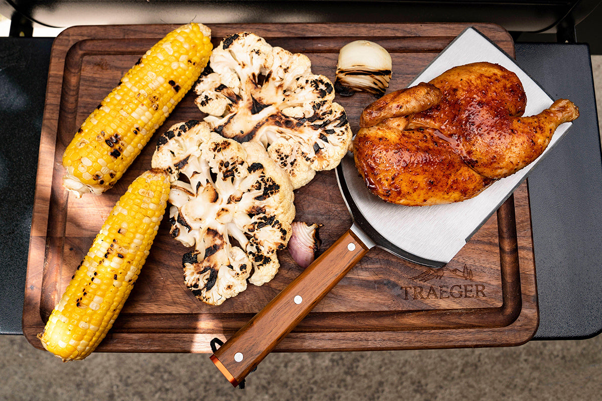 Traeger Large Cut BBQ Spatula holding cooked chicken beside grilled corn and cauliflower