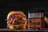 Delectable pulled meat sandwich with vibrant purple cabbage and orange carrot slaw, placed on a dark wooden surface. Adjacent to the sandwich is a container of Traeger Rub with labels highlighting its natural, gluten-free, and GMO-free qualities against a moody black backdrop.