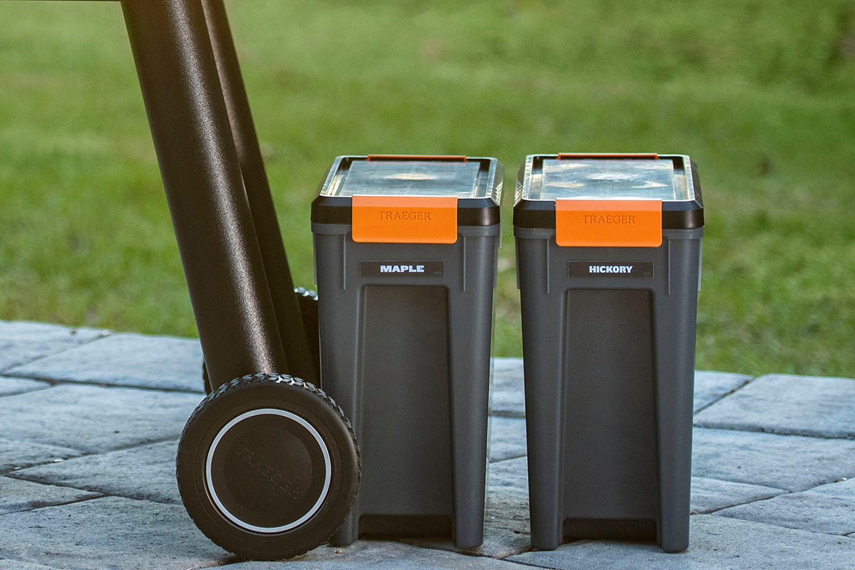 Two Traeger StayDRY pellet bins labelled Maple and Hickory beside a grill on a patio