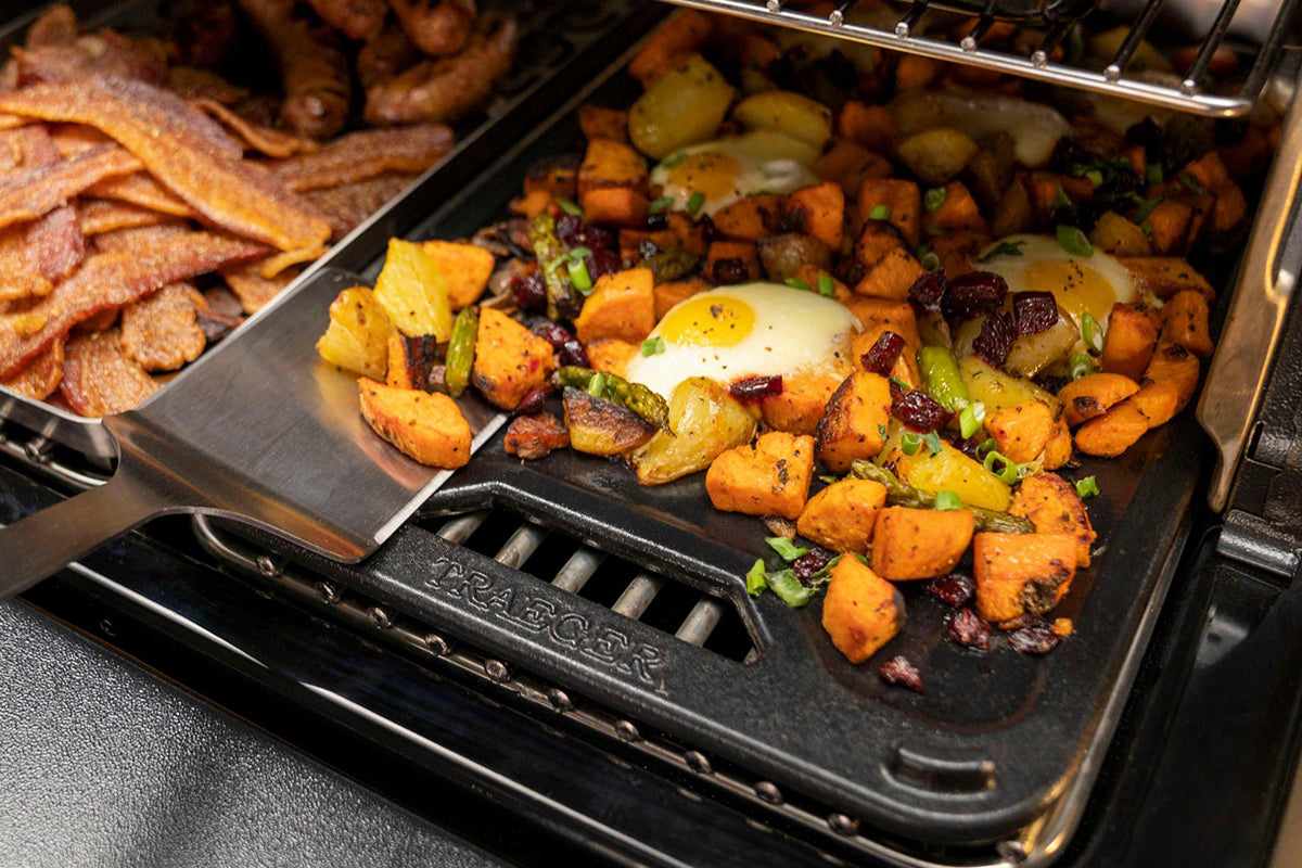 Eggs and diced vegetables cooking on the flat side of a Traeger griddle beside a tray of bacon