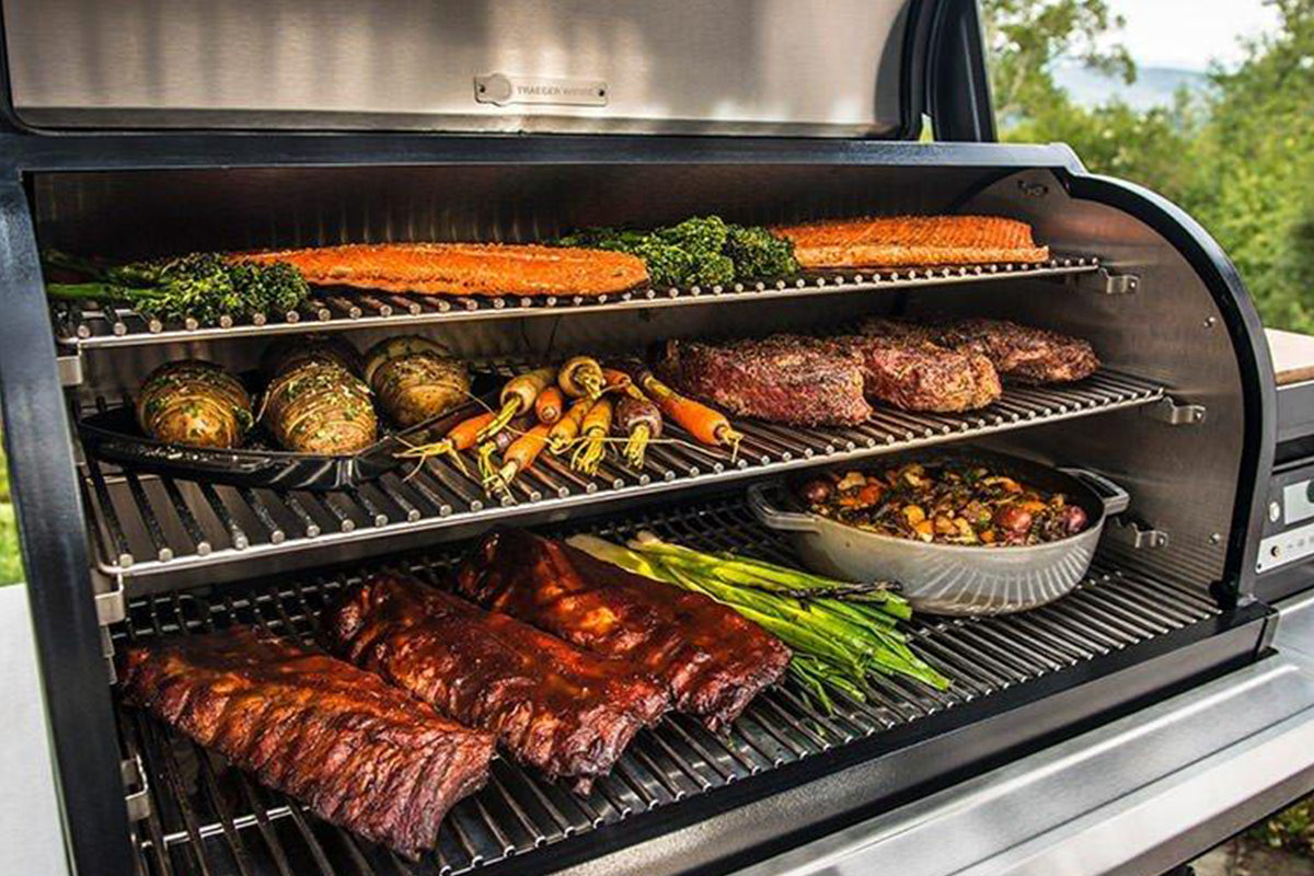 Open Traeger grill with three cooking racks holding salmon, meat and vegetables