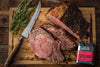 A sumptuously cooked prime rib, sliced to reveal its tender pink center, sits on a rustic wooden board. Accompanying it are sprigs of fresh rosemary and a vintage-styled knife with a droplet-laden blade, suggesting the freshness of the cut. To the side, a container of Traeger Prime Rib Rub underscores the dish&#39;s flavor profile. The ambiance of the image is enhanced by the wooden texture of the backdrop and scattered herbs and drippings.