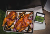 A seasoned slab of meat rests in a foil tray next to a black container of Traeger&#39;s &quot;Pork &amp; Poultry Rub&quot; with green labeling, highlighting its apple and honey flavor components.