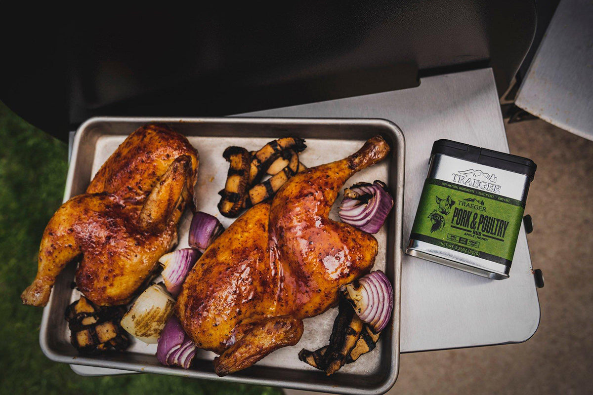 A seasoned slab of meat rests in a foil tray next to a black container of Traeger's "Pork &amp; Poultry Rub" with green labeling, highlighting its apple and honey flavor components.