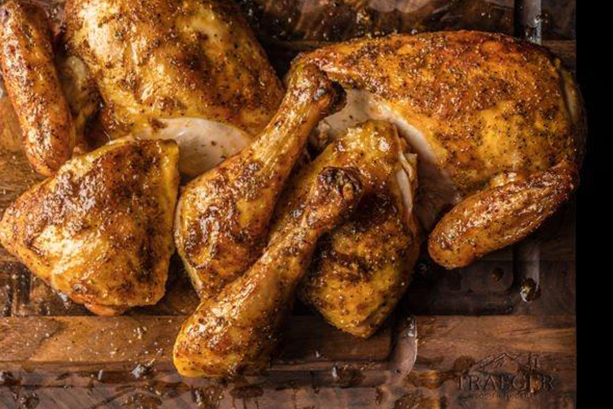A succulent roasted chicken, seasoned with a rich blend of spices, lies atop a rustic wooden surface. The golden-brown skin glistens with a savory glaze, and the chicken's tender meat hints at juicy flavors within. The embossed word "TRAEGAR" is faintly discernible on the wooden surface.