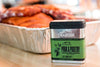 Seasoned meat in an aluminum tray next to a &quot;Traeger Pork &amp; Poultry Rub&quot; container, highlighting its 100% natural ingredients on a kitchen countertop.