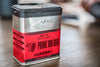 A Traeger Prime Rib Rub container placed on a wooden surface, captured in a shallow depth of field. The metal container has a black lid with visible perforations. The label is mainly red, showcasing the Traeger logo, an illustration of a bull&#39;s head, and the product name &quot;Prime Rib Rub&quot;. Some textual information is visible, including &quot;NET WT. 255g (9 oz)&quot;. The blurred background suggests an indoor setting with hints of other objects, though they are indistinct.