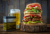 A tantalizing double-layered sandwich stacked with grilled meat, crispy bacon, fresh lettuce, tomato slices, and avocado on a rustic wooden slice. Beside it, a chilled glass of beer with a frothy top rests on a wooden table. To the left, a Traeger Fin &amp; Feather Rub tin prominently features its branding and imagery on a yellow label. The background is a weathered wooden wall.