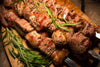 Grilled skewered meat with herbs, featuring succulent pieces wrapped in bacon, garnished with rosemary sprigs, on a rustic wooden surface.