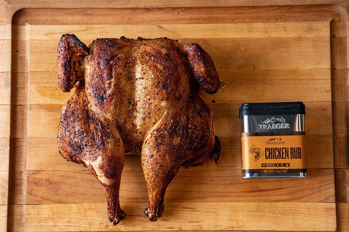 A perfectly roasted chicken with a crispy, seasoned skin is presented on a wooden cutting board. The rich brown hue of the chicken contrasts beautifully with the warm tones of the wood. Beside the chicken, there's a container labeled "TRAEGER CHICKEN RUB," indicating the seasoning used on the chicken. The container features the Traeger logo and specifies a net weight of 255g (9 oz).
