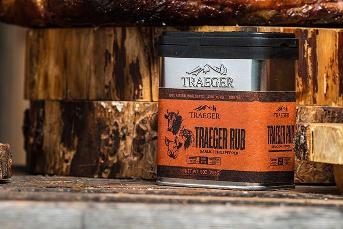 A container of 'Traeger Rub' with garlic and chili pepper flavors, set against a backdrop of weathered wooden logs. The seasoning's label highlights its 100% natural ingredients, gluten-free, and GMO-free attributes.